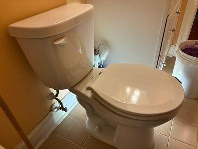 White toilet in a bathroom, with a closed lid. Adjacent to the toilet is a white wall, and a bucket is visible.