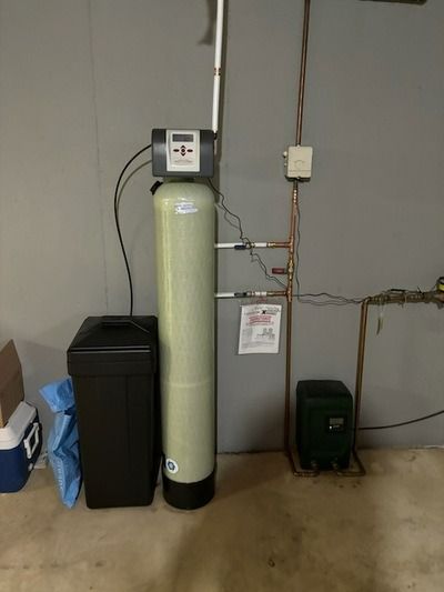 Water softener system in a basement setting. A tall beige tank, black brine tank, and green pump.