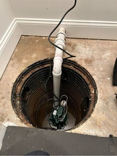 Sump pump in a pit, white PVC pipe, black electrical cord, concrete floor, ceiling trim visible.