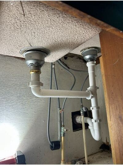 Underside of a kitchen sink with two drains connected by white PVC pipes. A cabinet door is visible on the right.