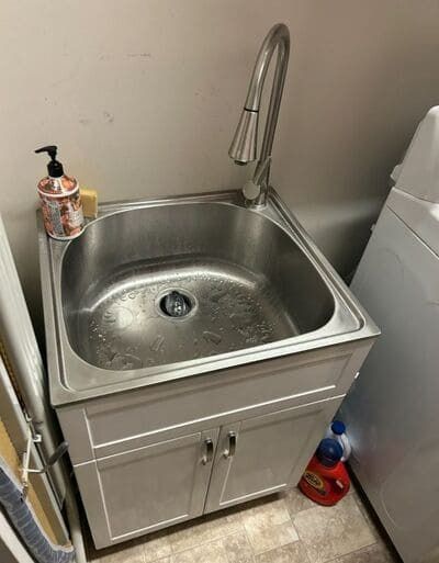 Stainless steel utility sink with cabinet, faucet, and soap dispenser in a laundry room.