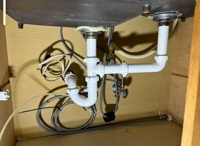 Under-sink plumbing with white pipes and a P-trap, connected to a dark sink bowl inside a wooden cabinet.