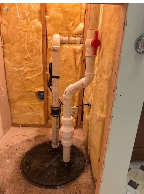 Sump pump installation in a framed wall with insulation, white PVC pipes, and a red valve.
