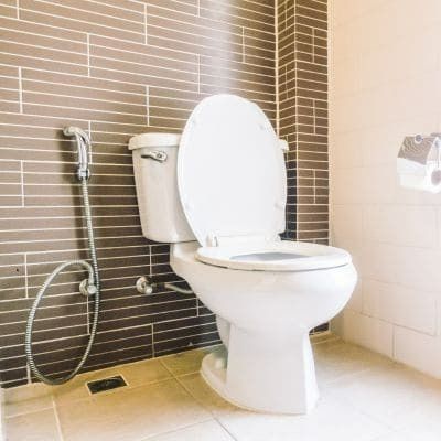 White toilet with the seat up in a bathroom with brown and beige tile walls. A bidet sprayer is attached.