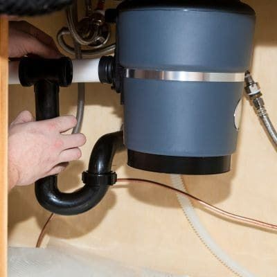 A person's hands connecting a black pipe to a gray garbage disposal under a sink.