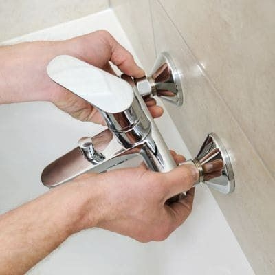 Hands installing a chrome faucet on a beige tiled wall.