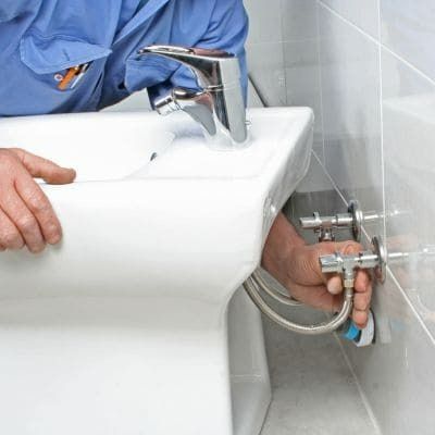 Plumber installing plumbing to a white bidet. Hands visible, working in a bathroom with white tile.