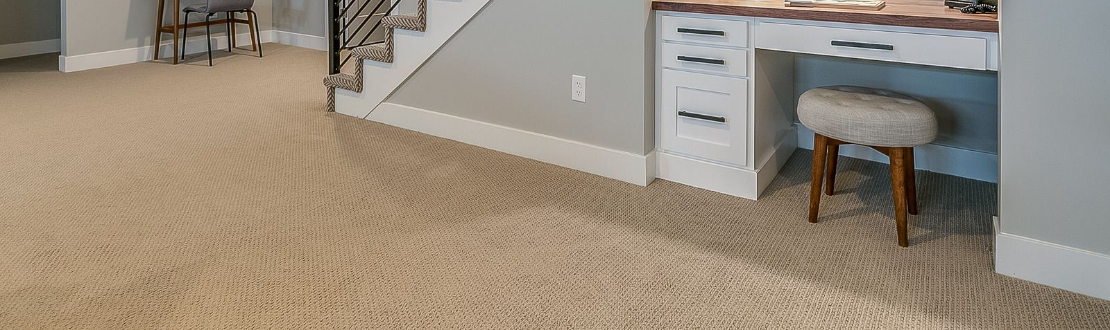 Beige carpeted room with a white desk and stool, a staircase, and chair.