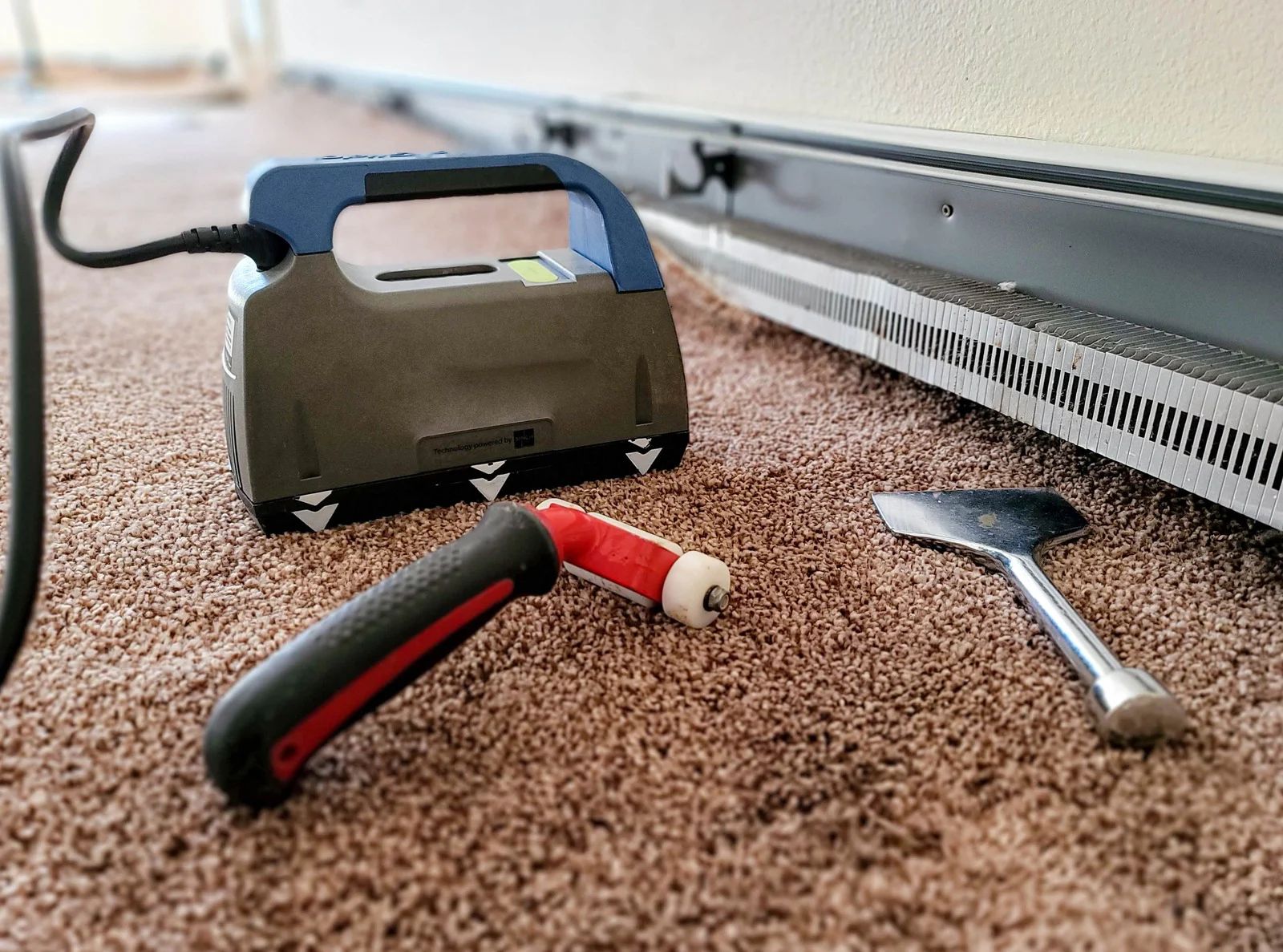 Carpet cleaning tools: machine, torch, scraper, on brown carpet next to a baseboard heater.
