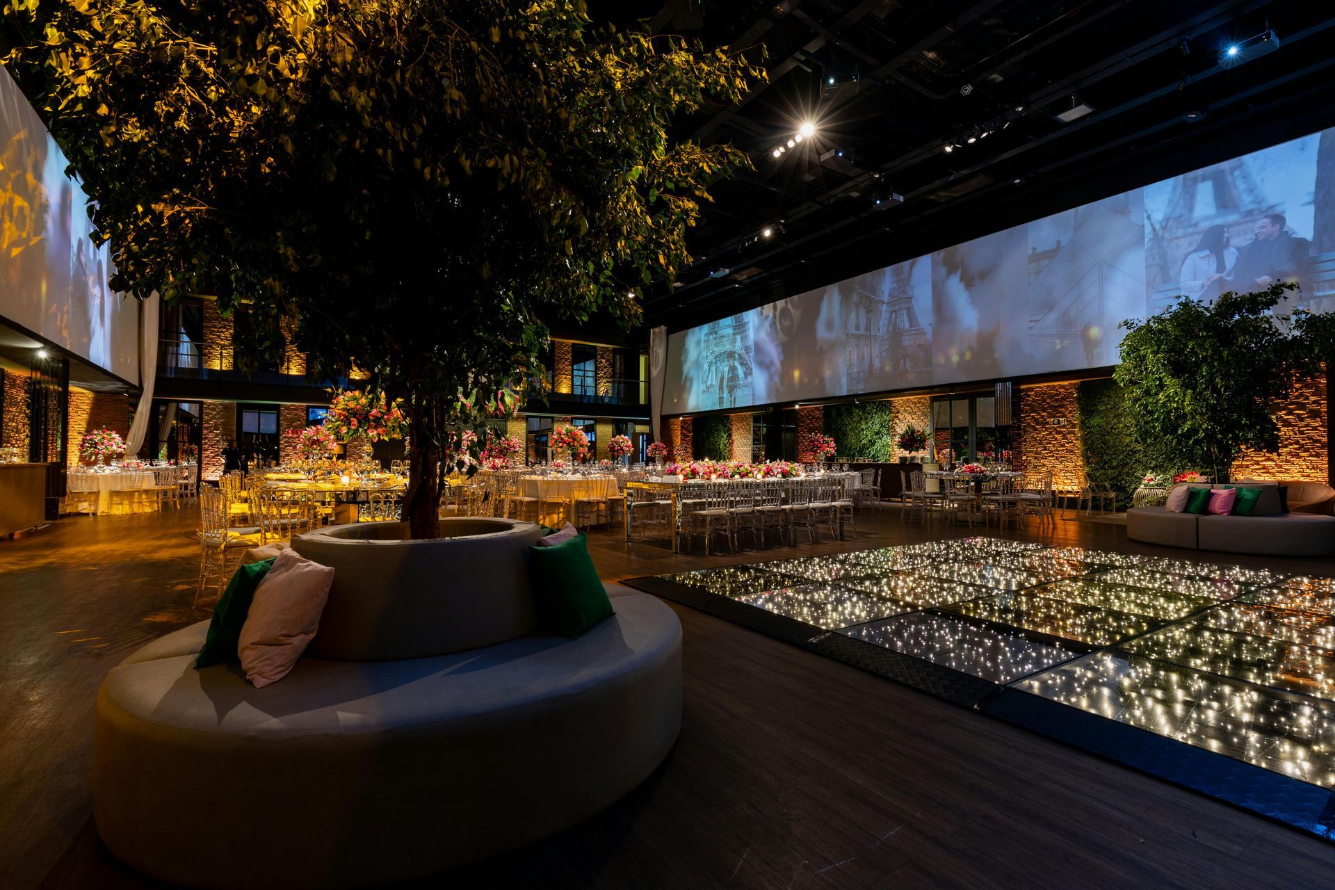 Indoor event space decorated for a celebration, featuring a round seating area, trees, tables, and a lit-up dance floor.