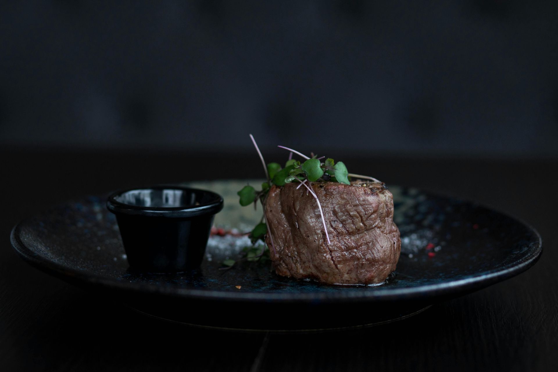 Close-up of a seared steak, garnished with greens, on a black plate with a small dish of sauce. Dark background.