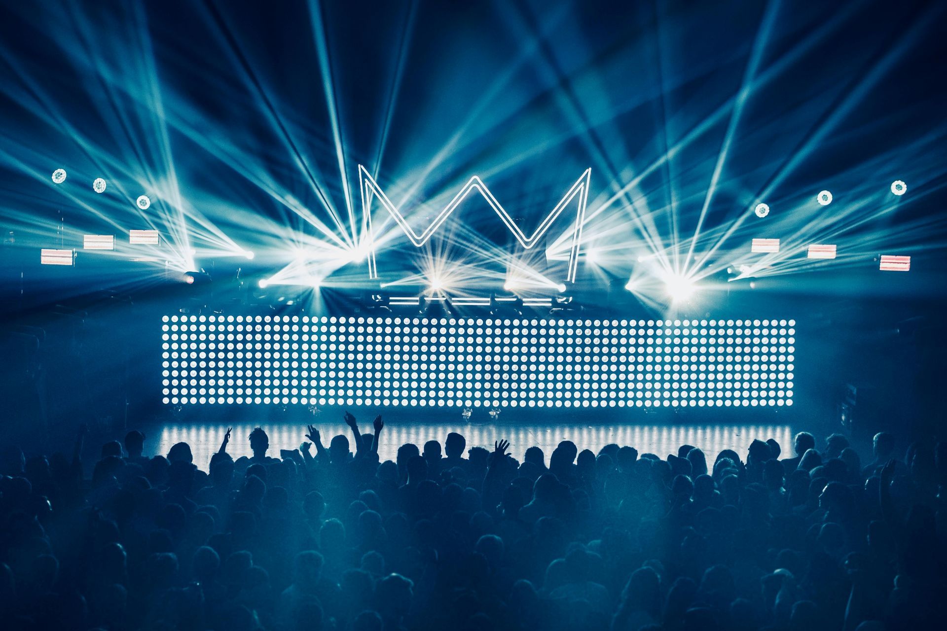 Concert stage with bright blue lights shining on a crowd, and a digital screen shaped like a crown above the DJ booth.