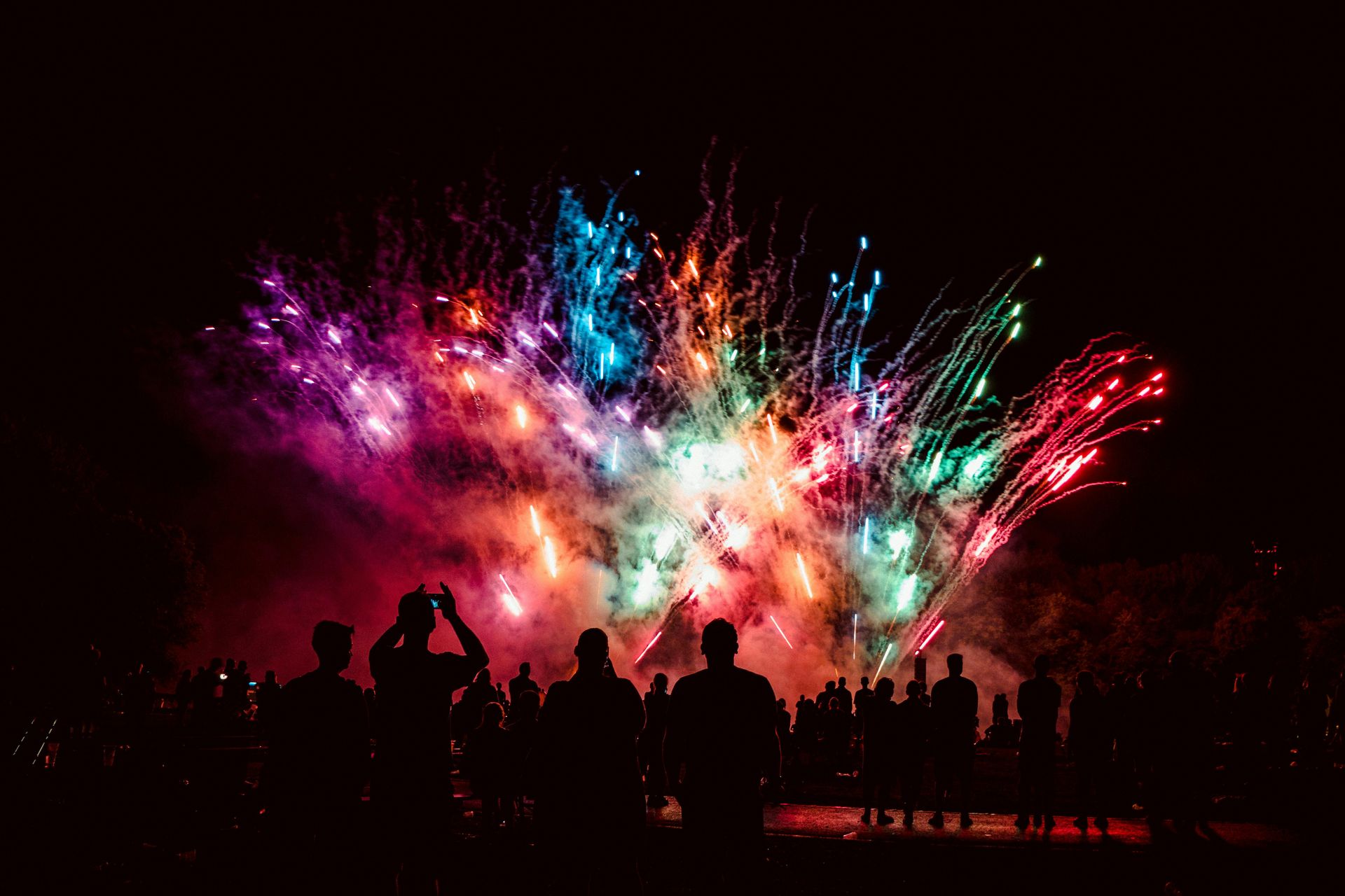 Fireworks explode in the night sky above a silhouetted crowd, displaying vibrant bursts of color.