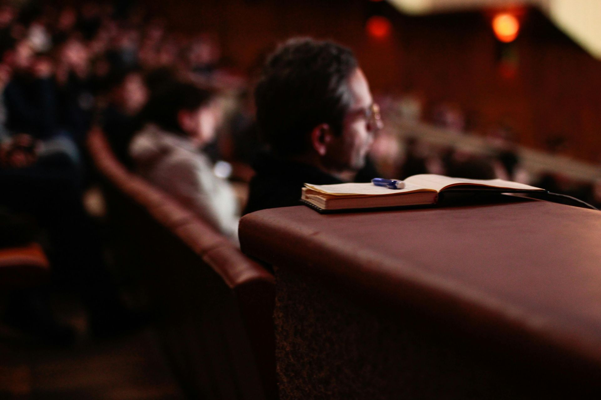 A man in glasses sits in a theater, a notebook open on the armrest in front of him. People fill the seats.