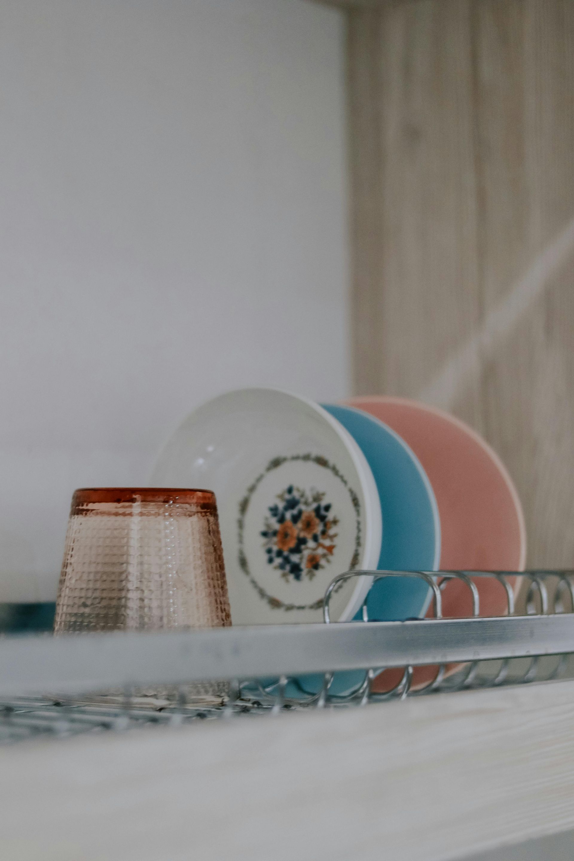 A dish rack holds a glass and a stack of plates. The plates are white with a floral design, blue, and pink.