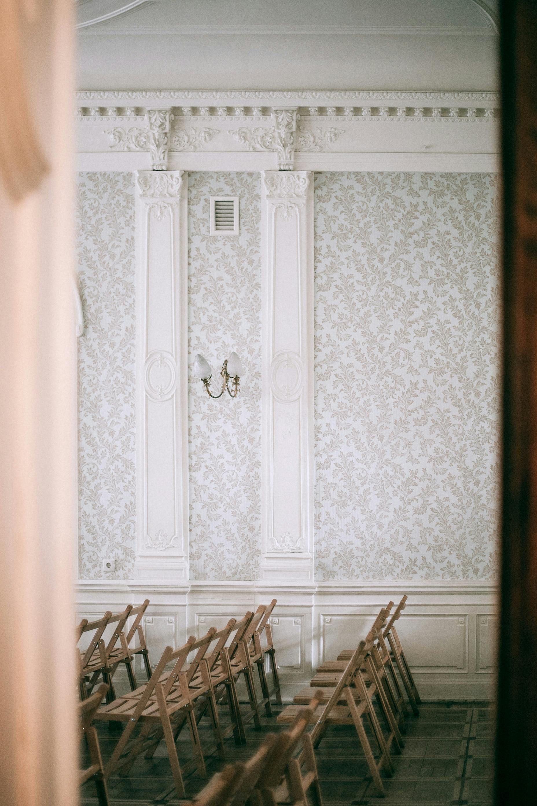 A row of wooden folding chairs faces a patterned wallpapered wall, framed by a doorway. A sconce hangs on the wall.