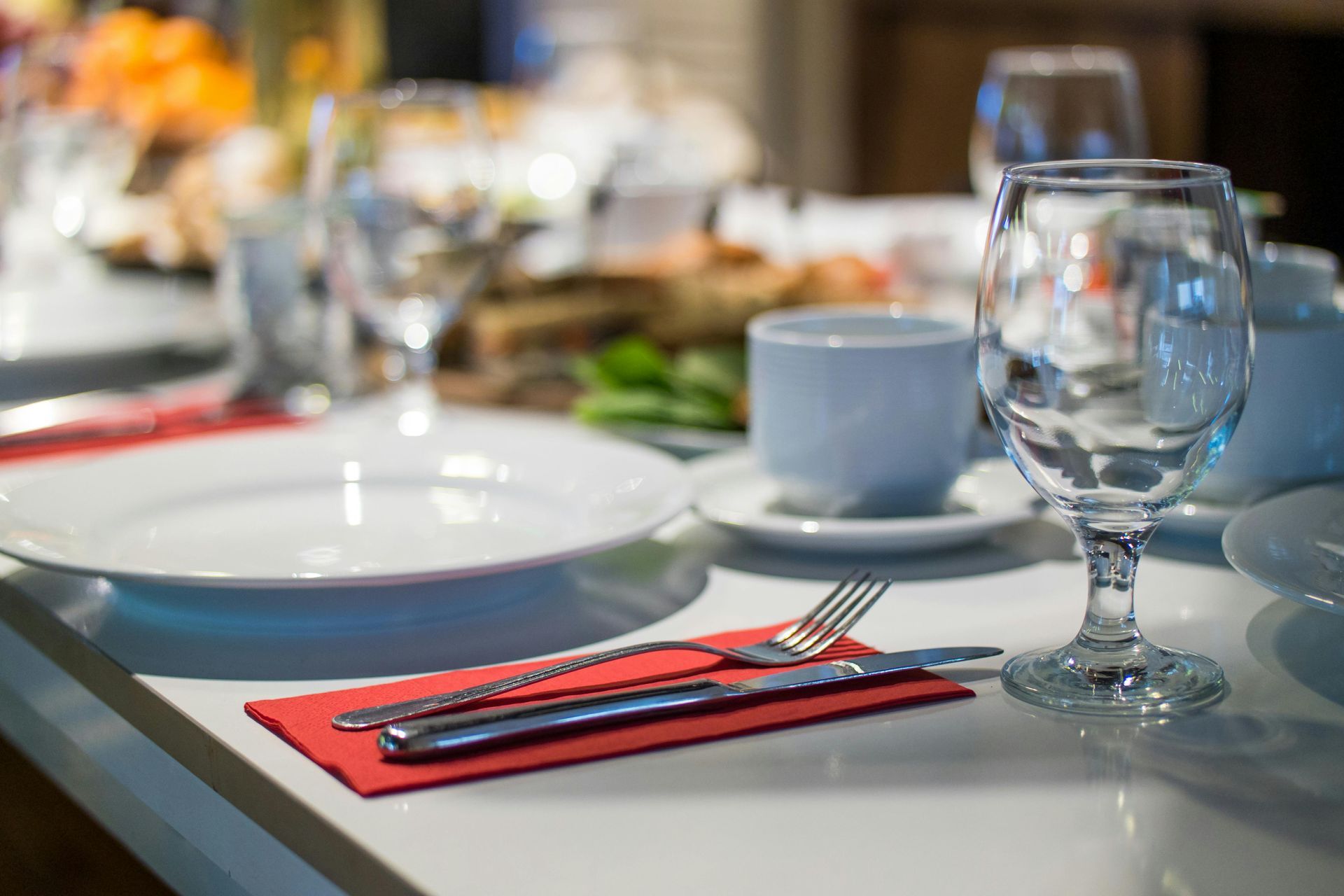 Table setting: White plate, silverware on red napkin, water glass, blue teacup, blurred food items.