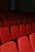 Rows of empty red theater seats fill the frame, gradually darkening towards the back, with a glimpse of a wall in the upper right.