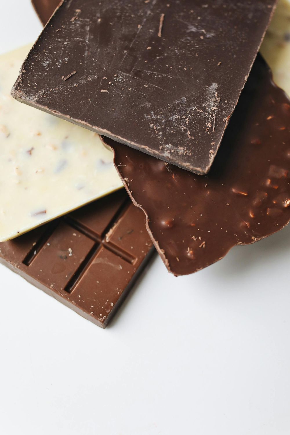 Vari pezzi di cioccolato, tra cui fondente, al latte e bianco, su una superficie bianca.