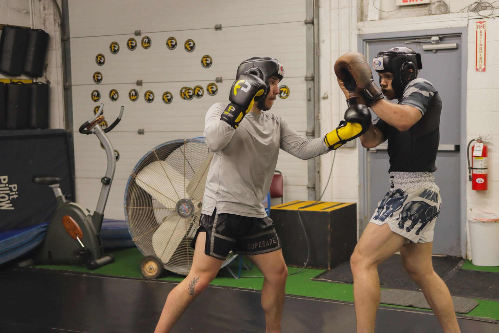 Two men are boxing in a gym with a fan in the background.