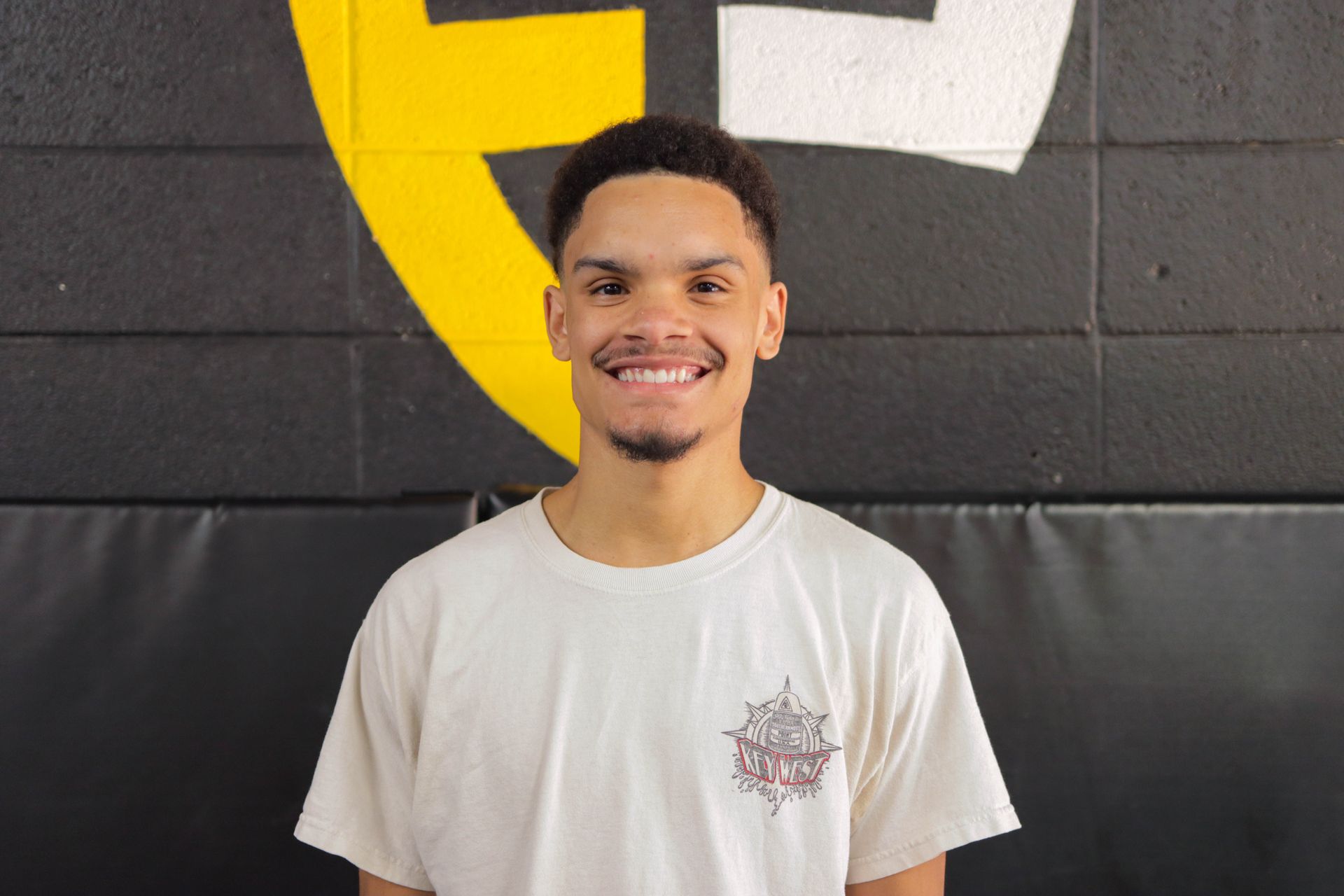 A man in a white shirt is smiling in front of a wall with a yellow and white circle on it.