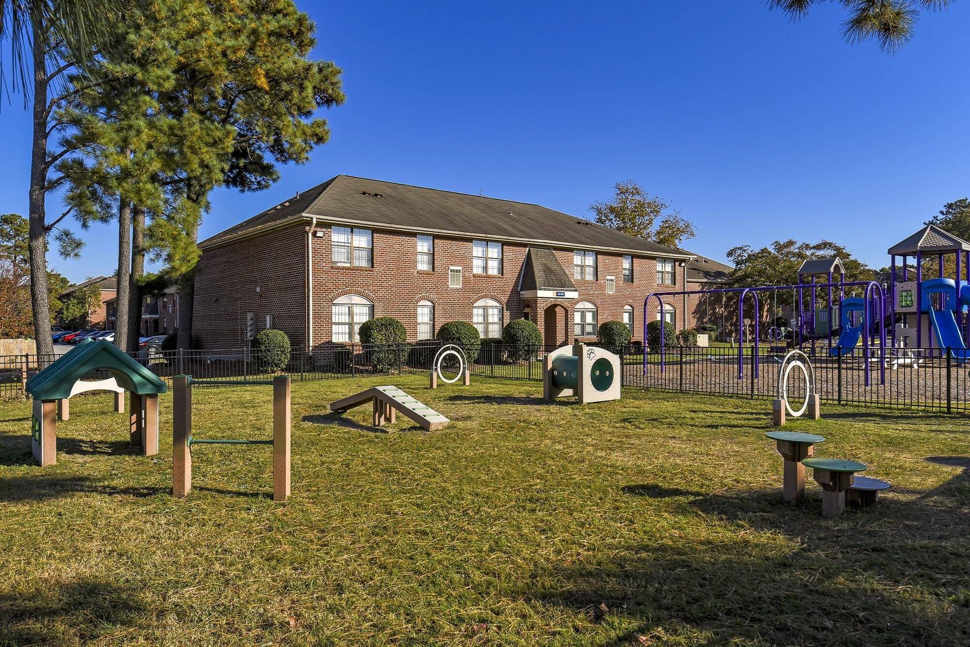 Brick apartment building behind a fenced playground with colorful play structures on a sunny day.
