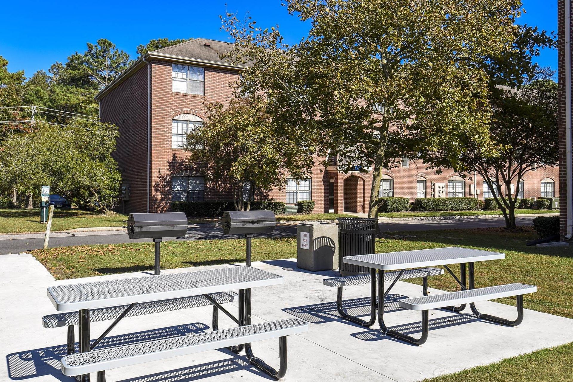 Outdoor communal picnic area with metal tables, grills, and trees near brick apartment buildings.