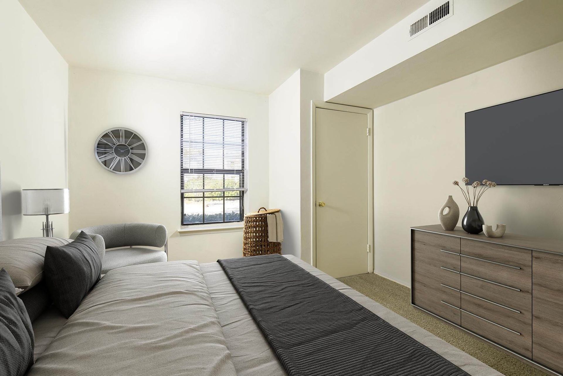 Bedroom in an apartment with a bed, dresser, wall clock, and a window.