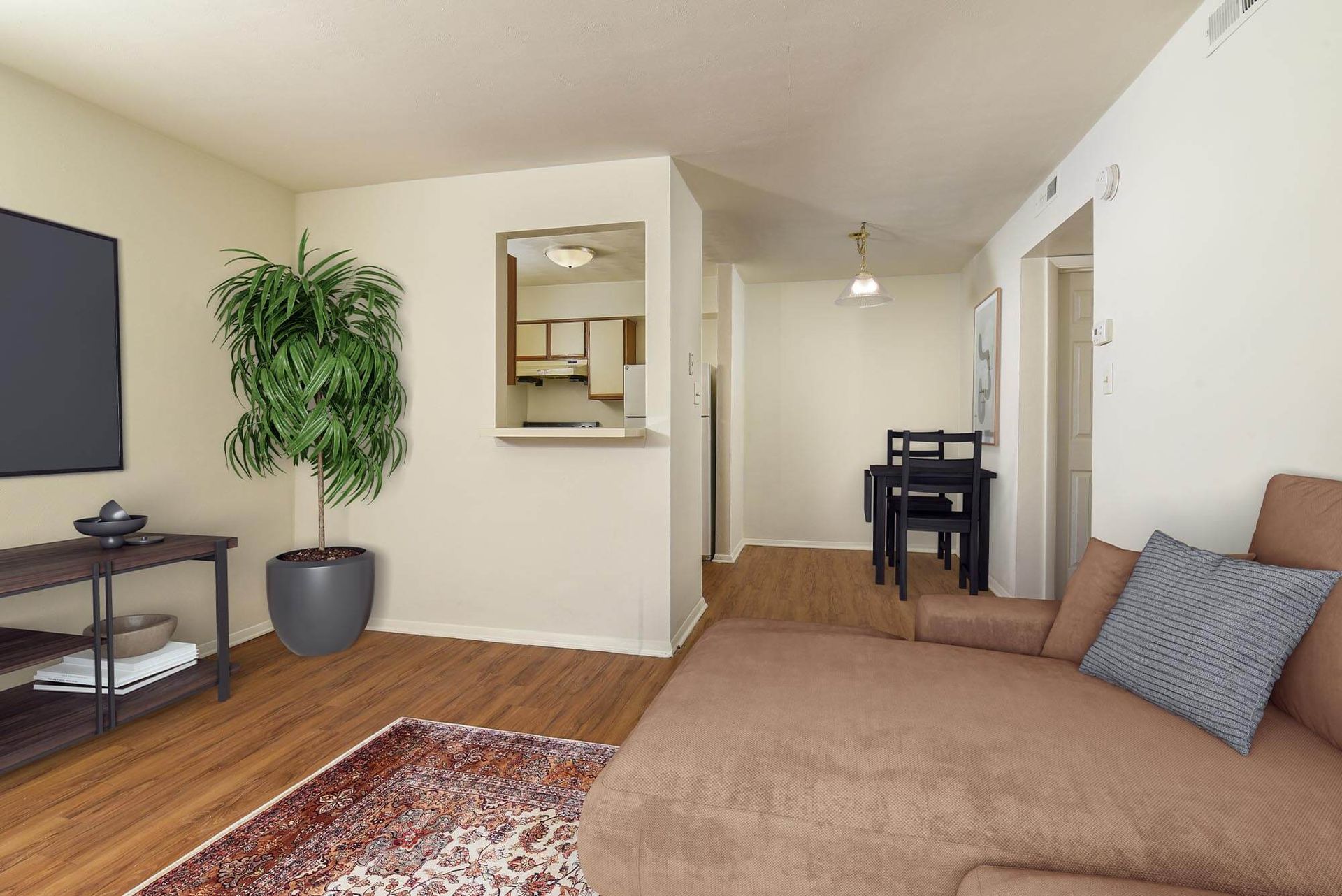 Living room with a brown sectional sofa, flat-screen TV, tall plant, and view into the open kitchen.