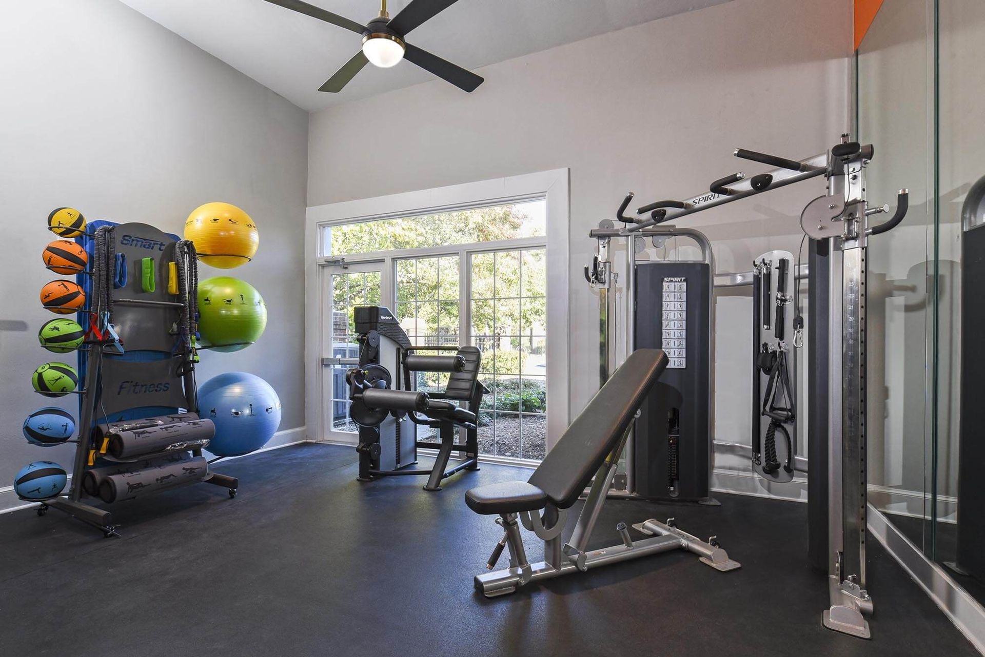 Interior fitness room with a multi-gym, weight bench, and colorful medicine balls near a window.