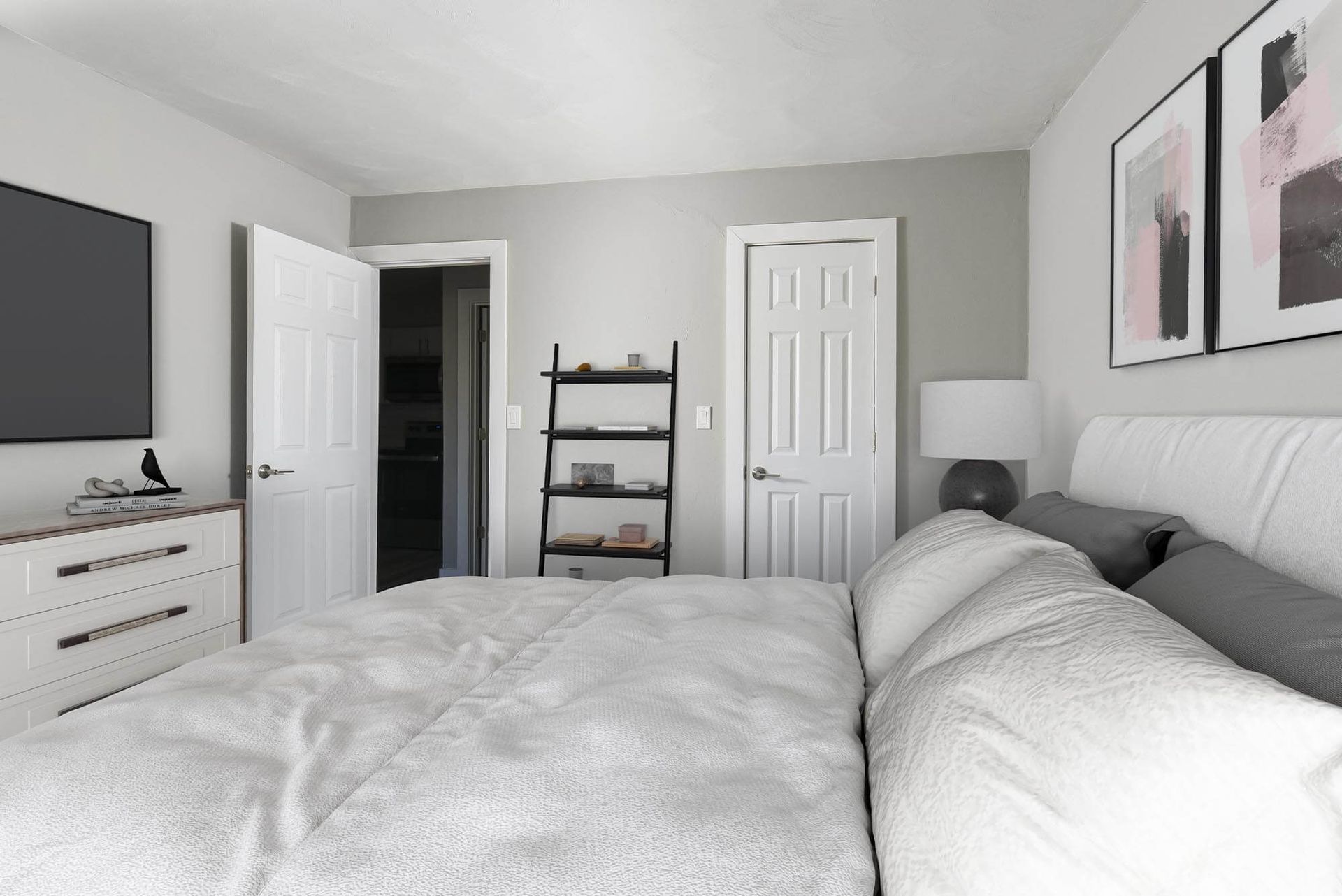 Bedroom interior with a large bed, white dresser, wall TV, bookshelf, and two doors.