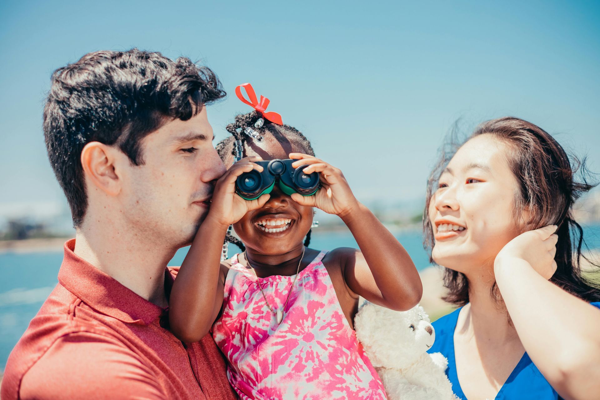 Family of three outdoors, little girl using binoculars, smiles.
