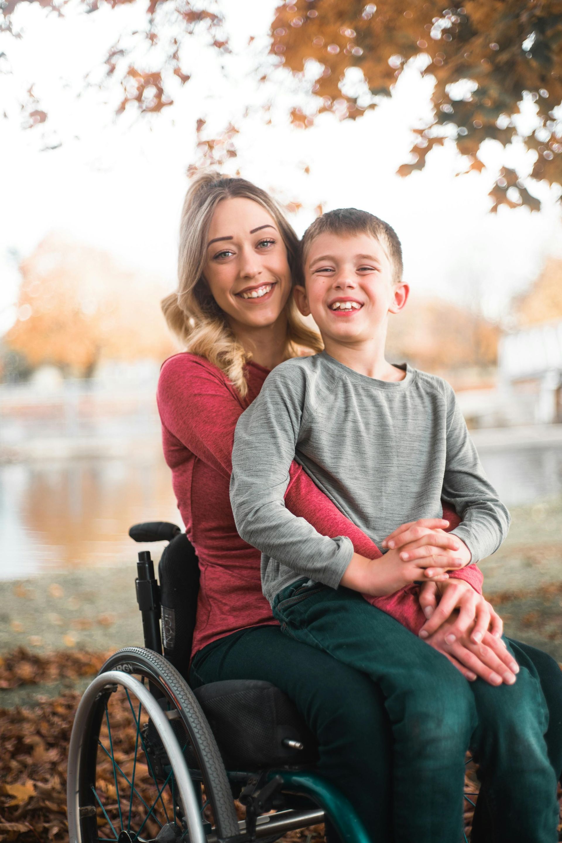 Woman in wheelchair smiling, holding a smiling child, in front of a pond and fall foliage.