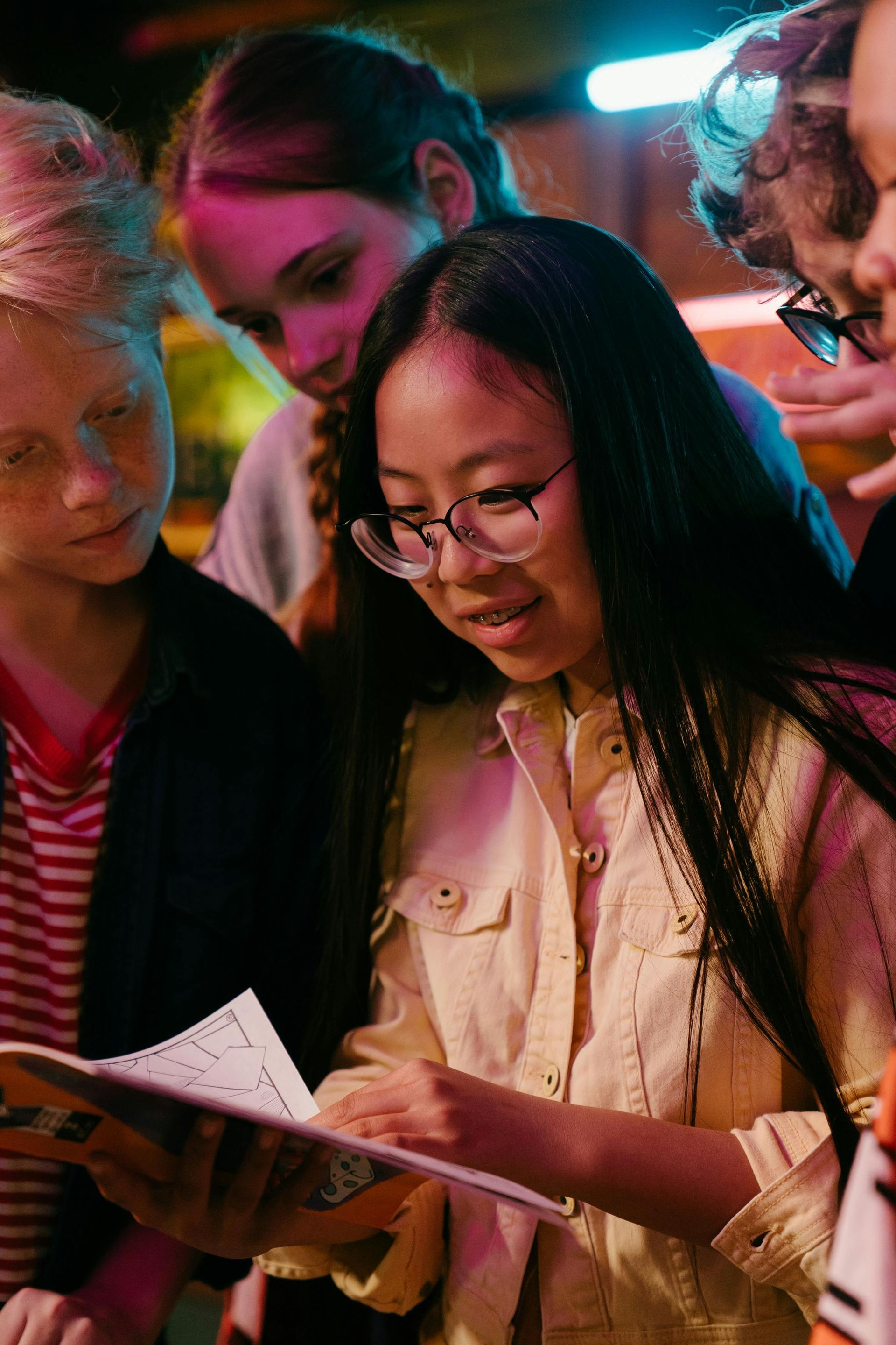 Girl with glasses reading a book, surrounded by other children, indoors, looking intently.