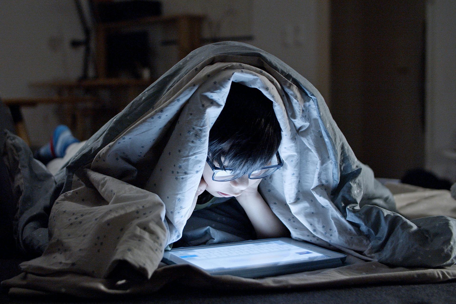 A tween wearing glasses under a blanket, illuminated by a tablet screen in a dimly lit room.