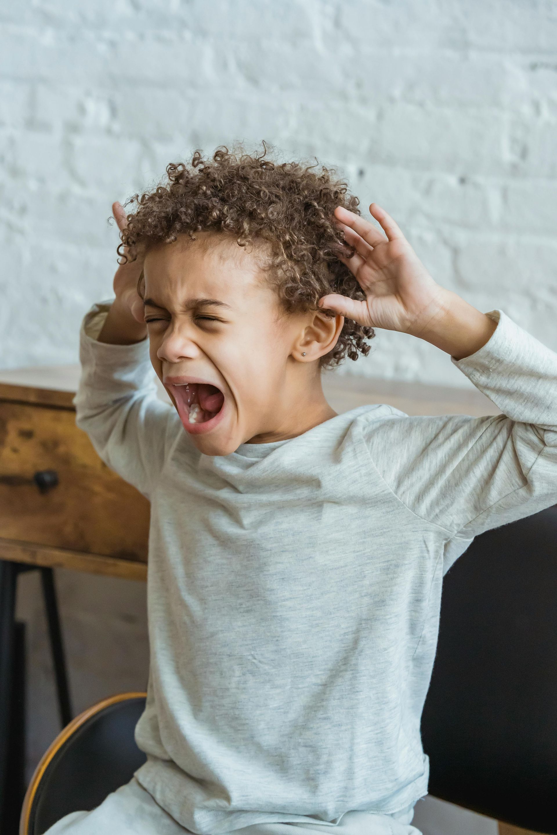 Young Black child with curly hair, screaming with hands up to head, seated indoors.