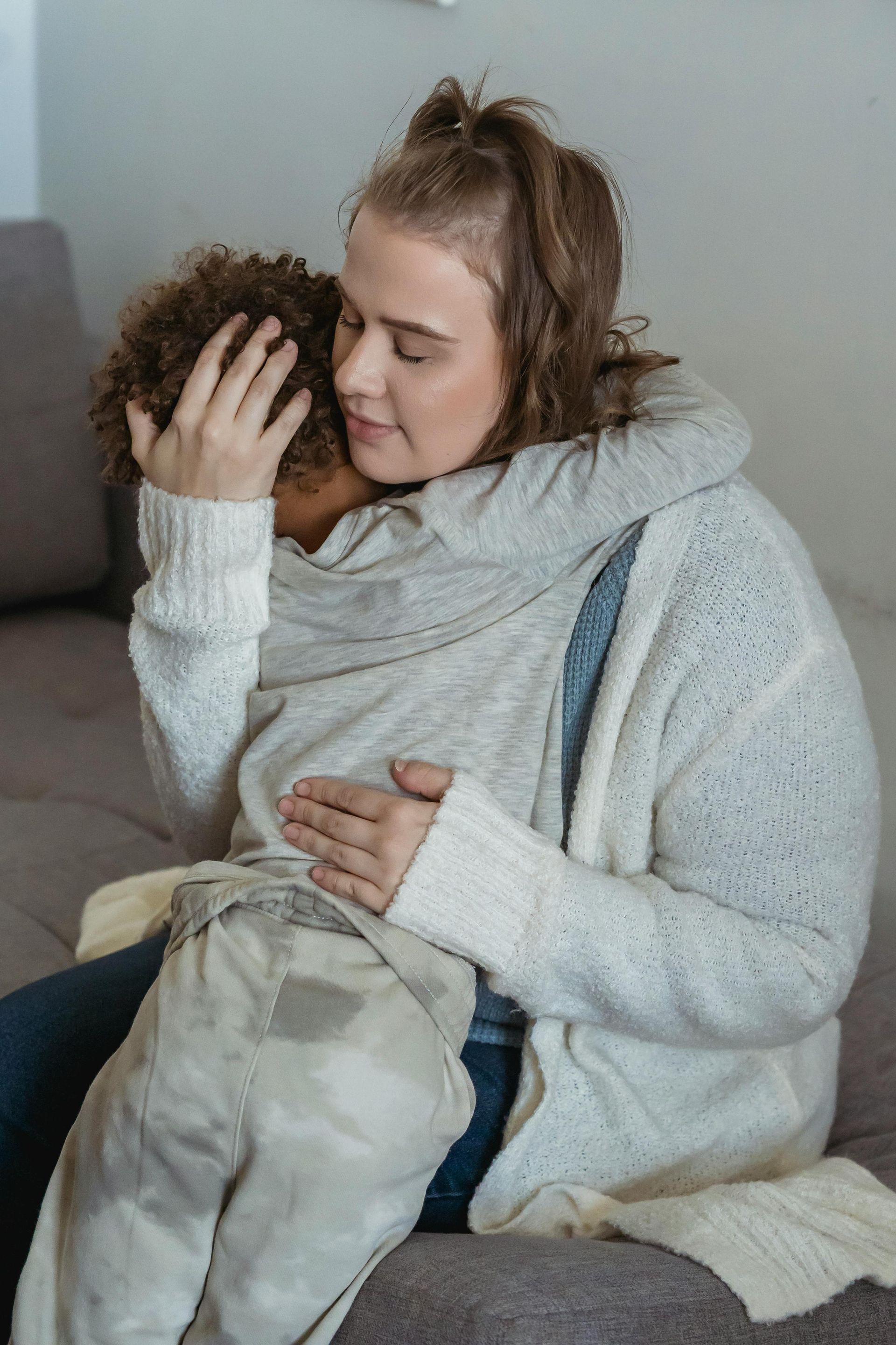 Woman embraces child, both seated on a couch. The woman is wearing a white cardigan.