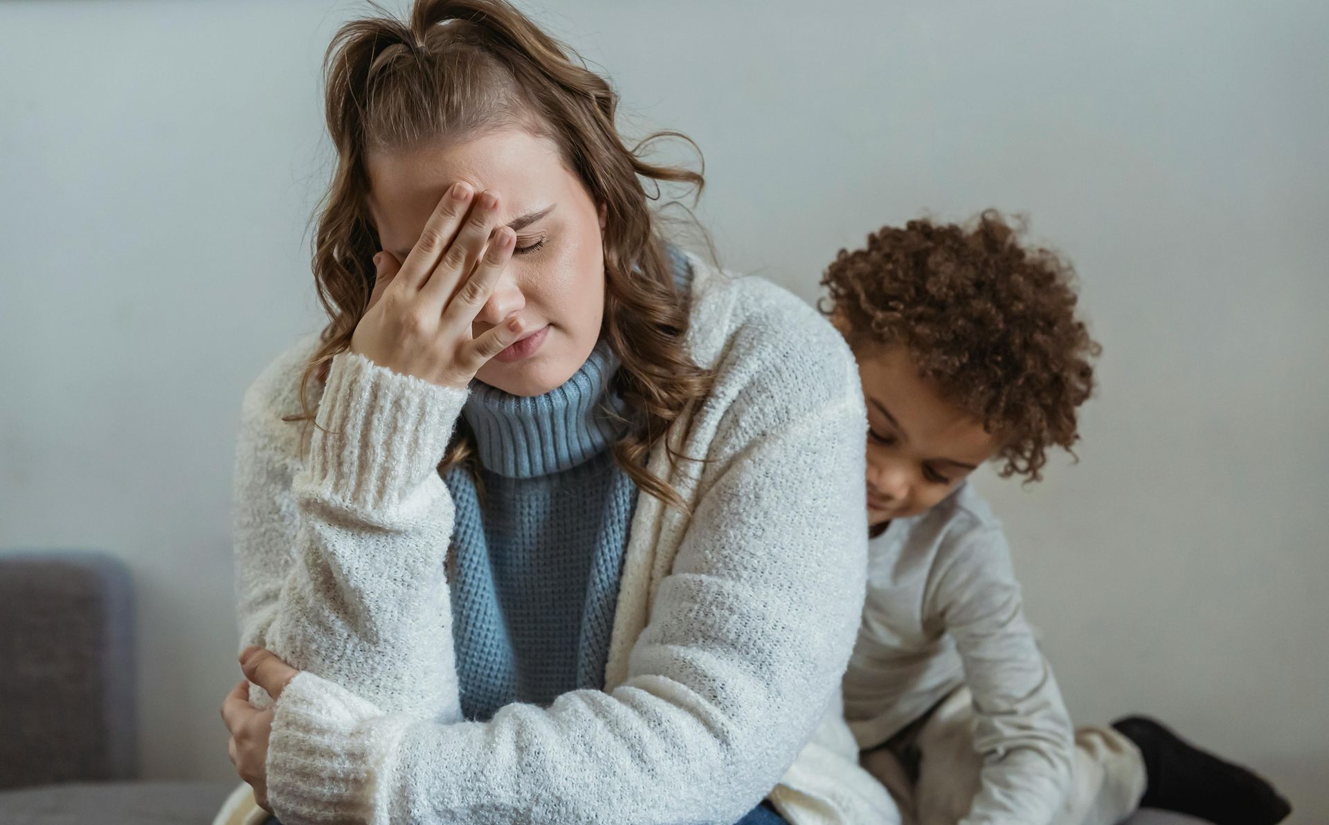 Woman with head in hand, looking distressed, while child hugs her from behind. Indoor, neutral colors.