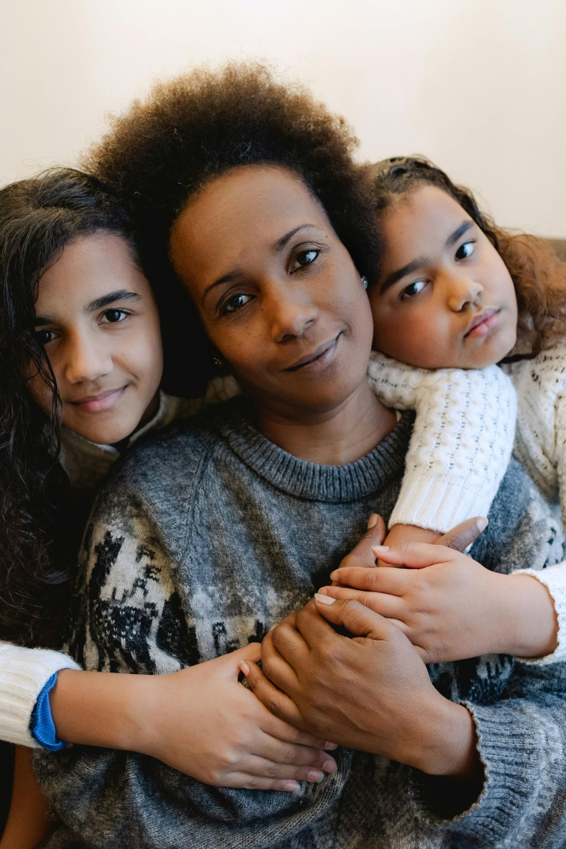 Mother and two daughters embrace; smiling woman, girls in sweaters, light background.