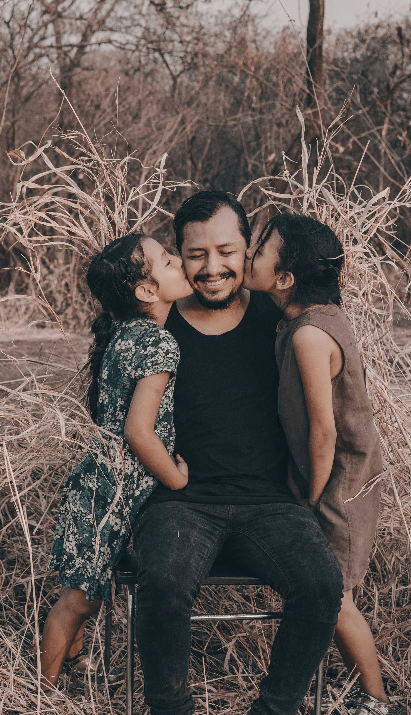 Man sitting, kissed on each cheek by two young girls, smiling outdoors.