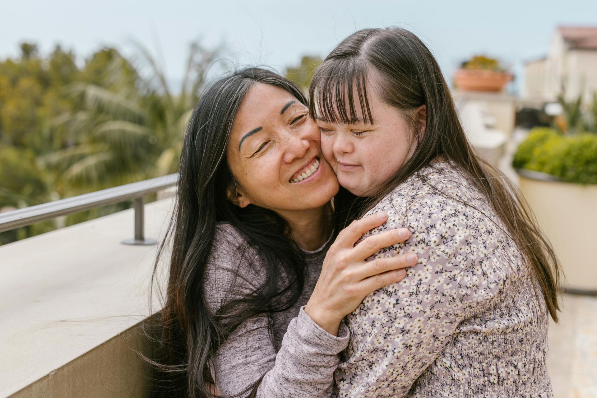 Woman embraces a person with Down syndrome; both smile, outdoors on a balcony.