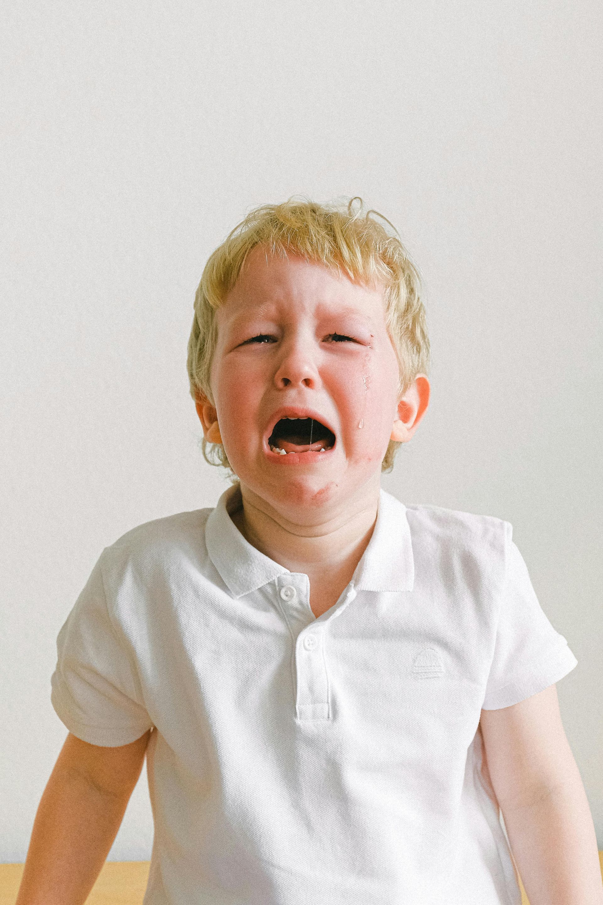A young child, with blonde hair and a white shirt, crying with mouth open, eyes closed.