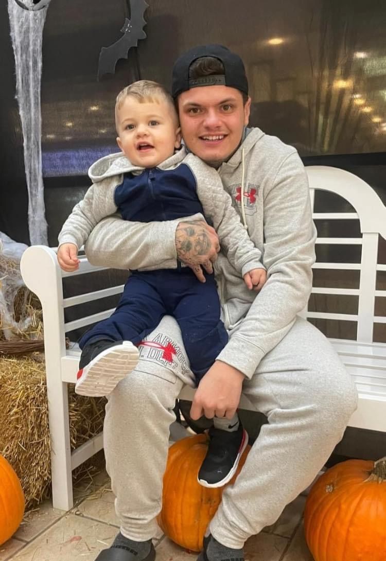 A man is holding a baby while sitting on a bench surrounded by pumpkins.