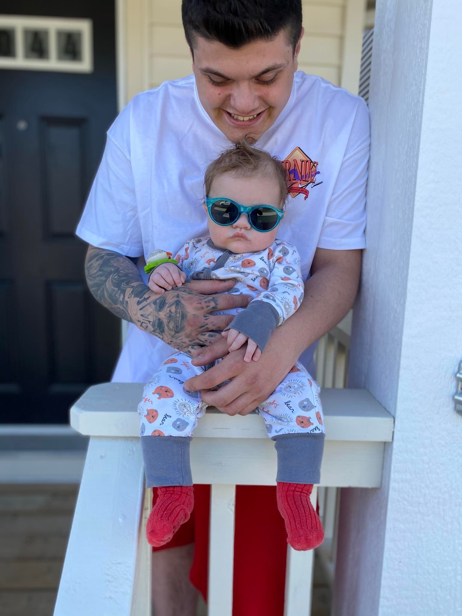 A man is holding a baby wearing sunglasses on a porch.