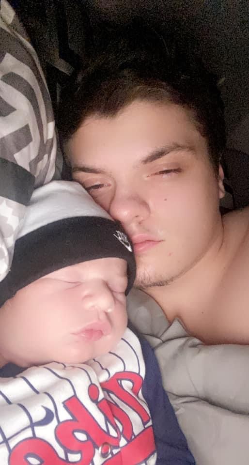 A man is sleeping next to a baby wearing a baseball cap.