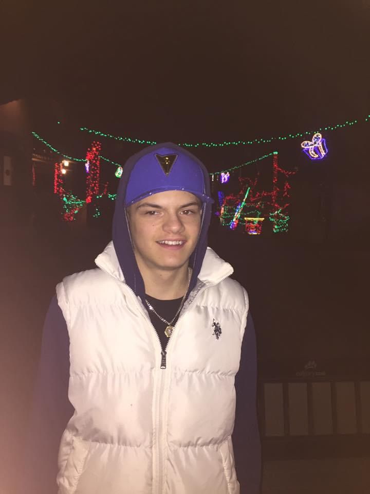 A young man wearing a purple hat and a white vest is standing in front of christmas lights.