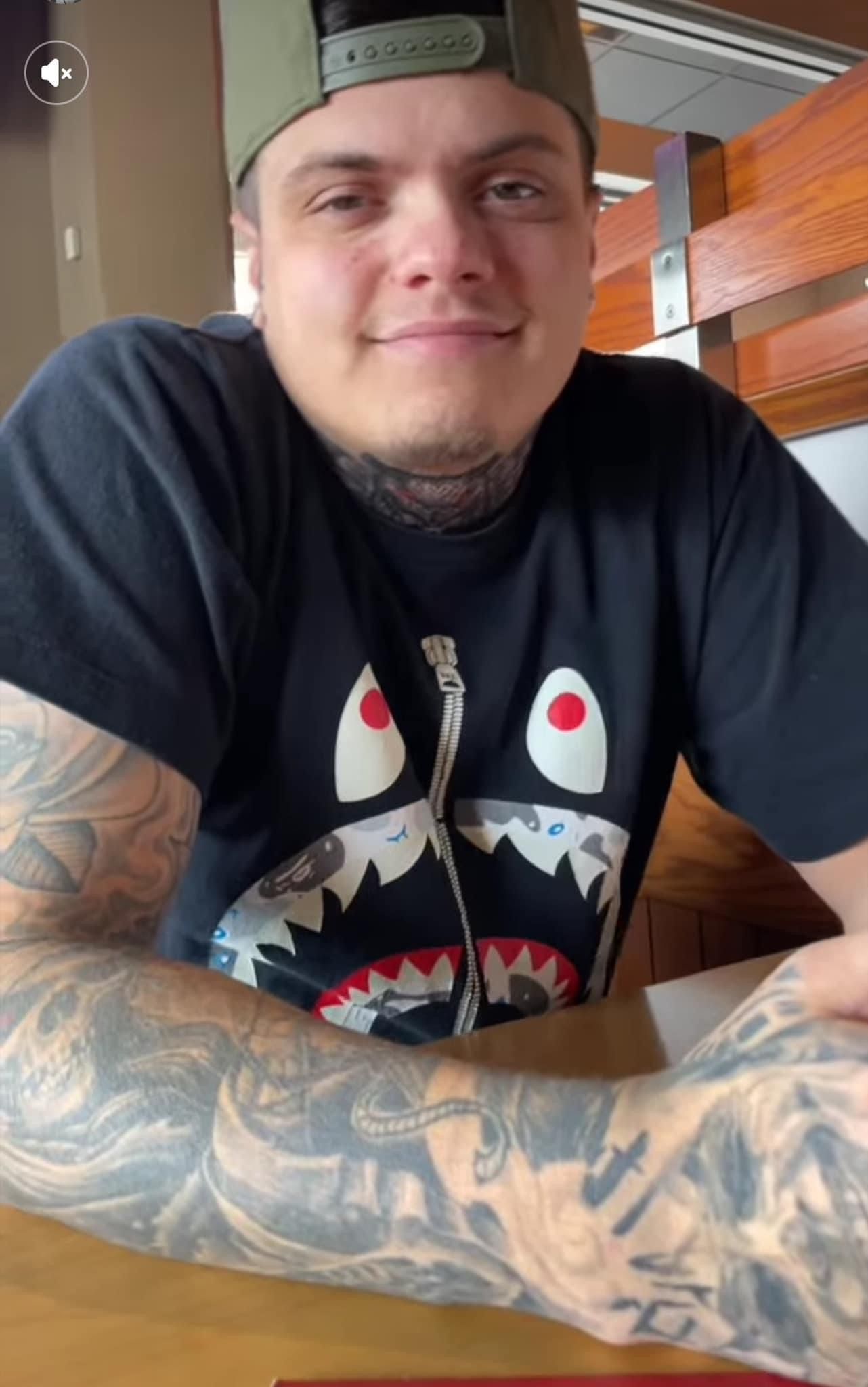 A man with tattoos is sitting at a table wearing a shark shirt.