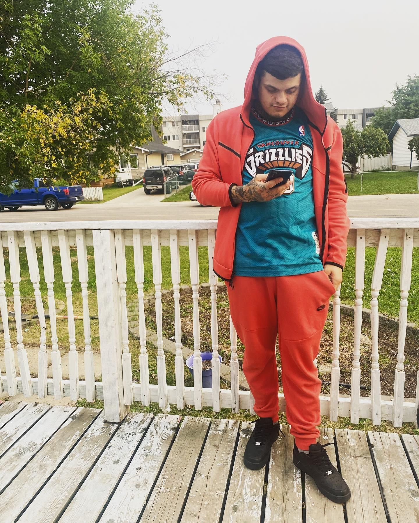 A man wearing a grizzlies shirt is standing on a deck looking at his phone.