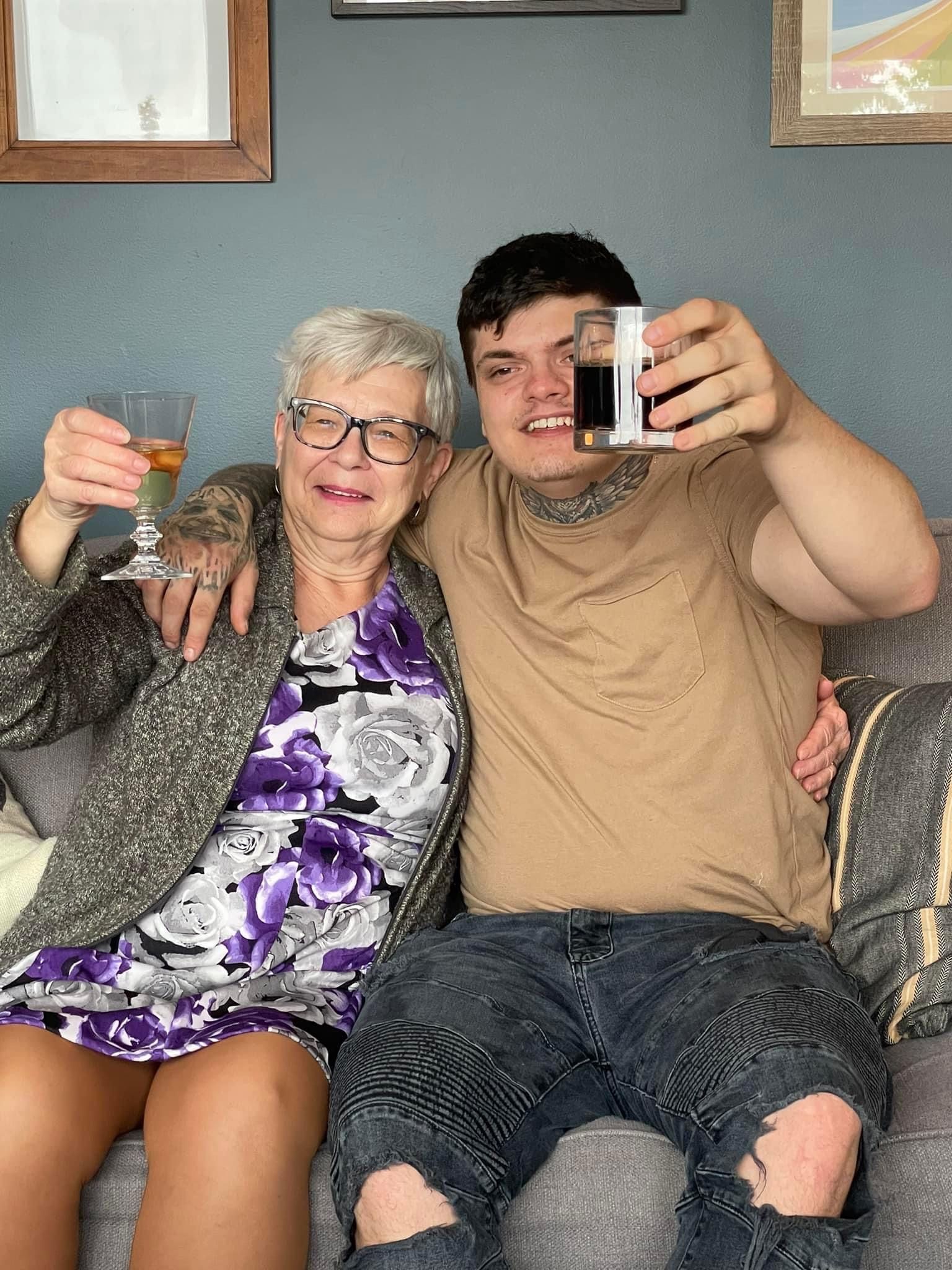 An elderly woman is sitting on a couch next to a young man holding a glass of wine.