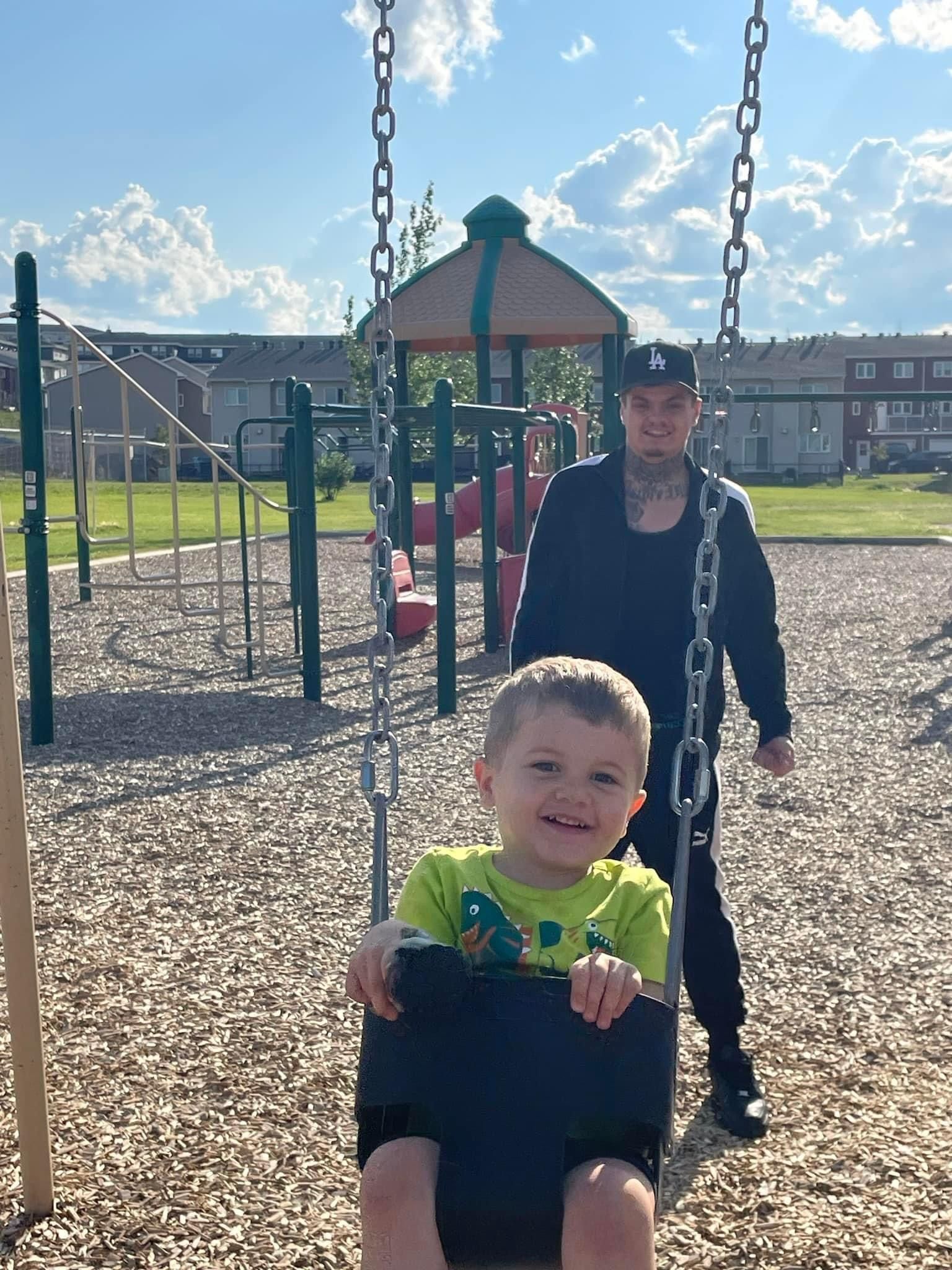 A little boy is sitting on a swing in a park while a man stands behind him.